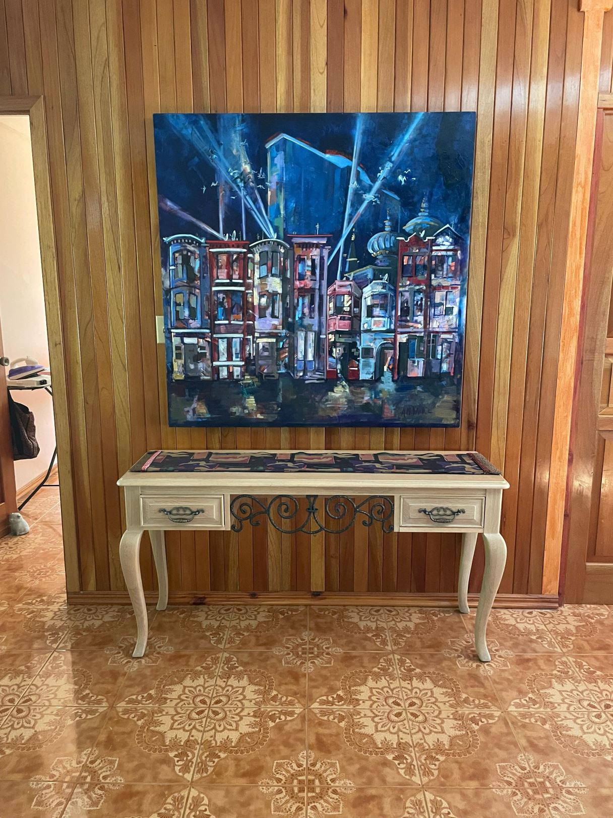 Abstract cityscape painting in blue tones mounted on a wood-paneled wall above a white console table with decorative metalwork.