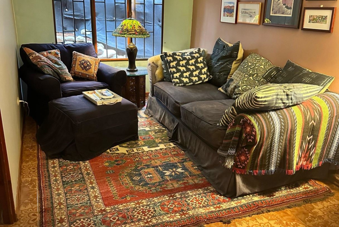Cozy living room corner with a dark sofa and armchair, colorful throw pillows, a Tiffany-style lamp, and a multicolored rug beneath.