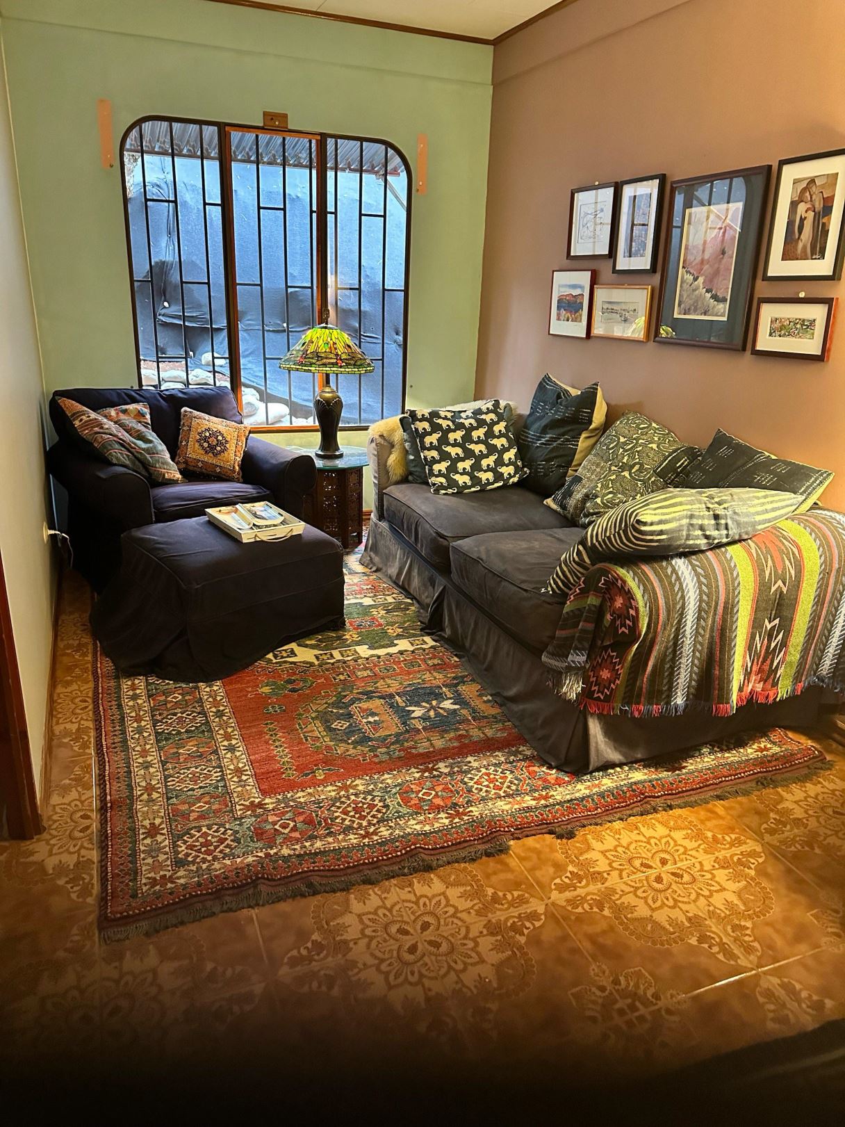 Cozy living room corner with a dark sofa and armchair, colorful throw pillows, a Tiffany-style lamp, and a multicolored rug beneath.