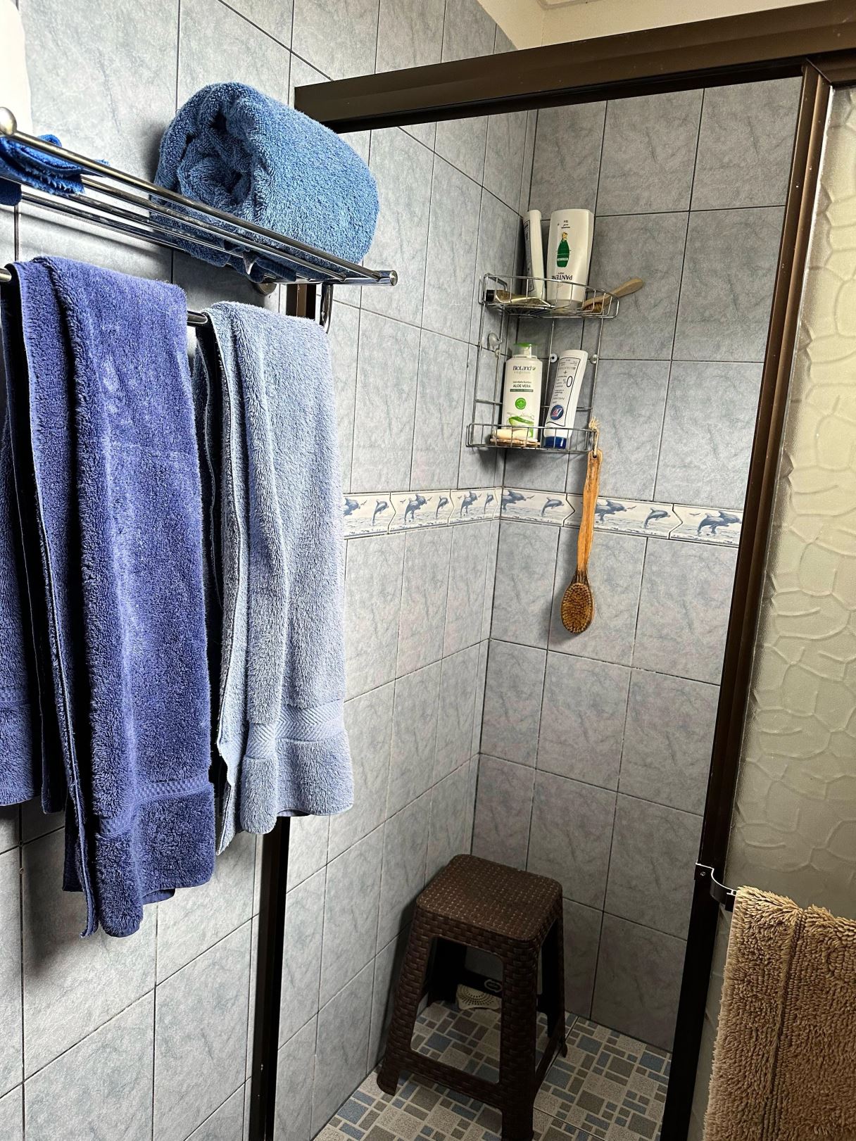 Blue towels hanging on a chrome rack in a blue-tiled bathroom with a small wicker stool beneath.