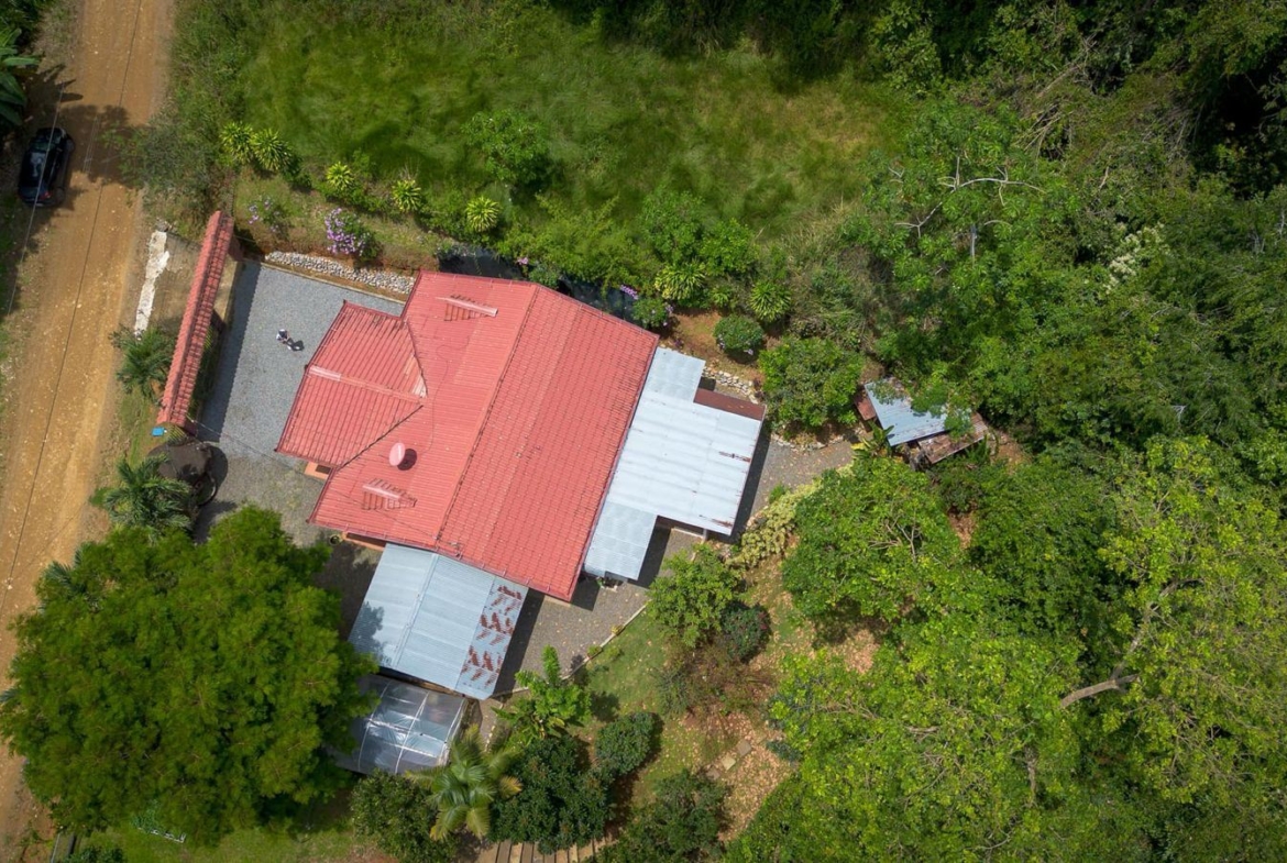 Aerial view of a red-roofed house with metal extensions, surrounded by trees and a dirt road on the left.