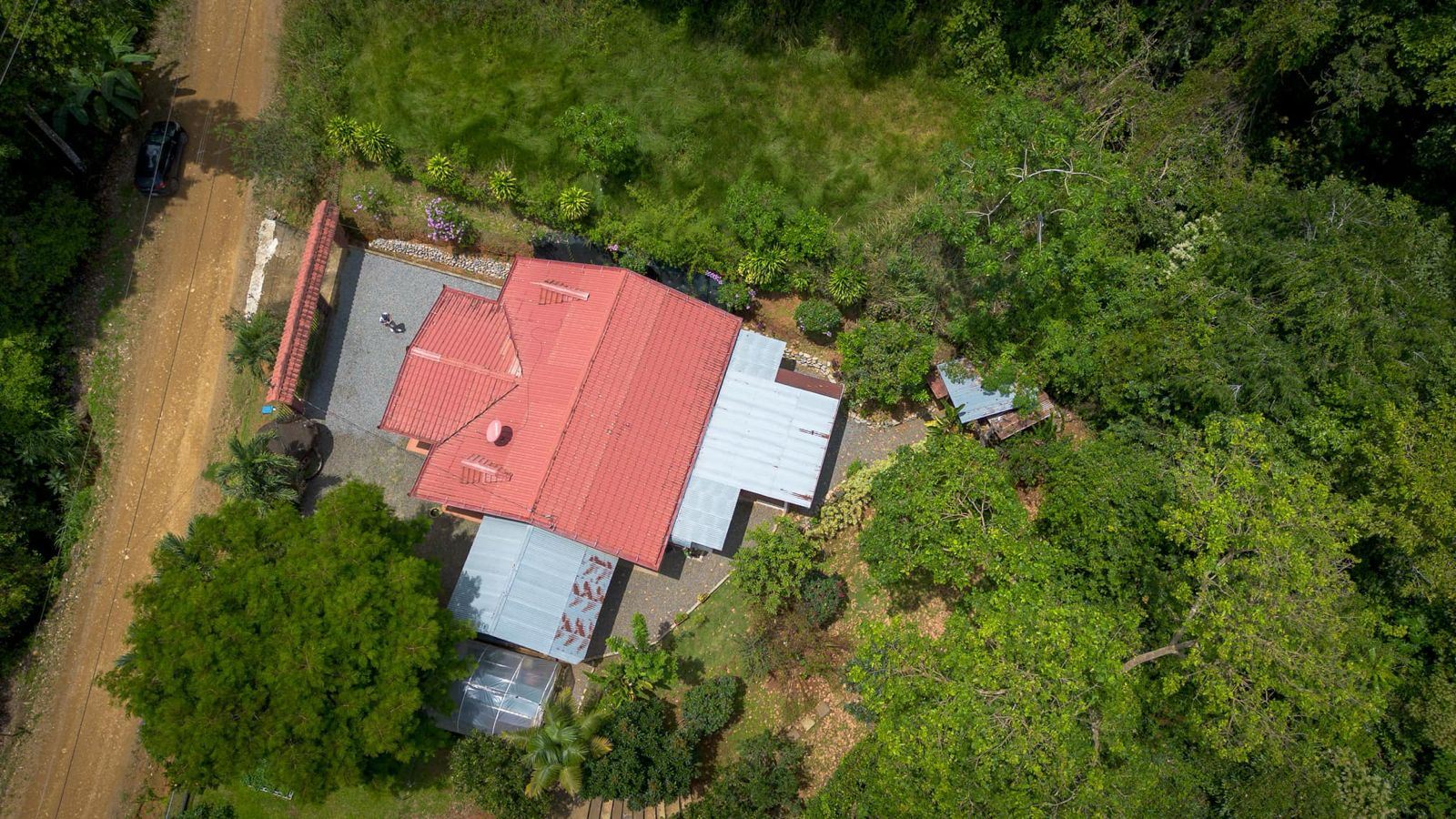 Aerial view of a red-roofed house with metal extensions, surrounded by trees and a dirt road on the left.