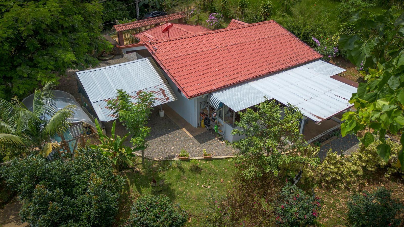 Aerial view of a house with a red tiled roof, metal awnings, and surrounding greenery in a garden setting.