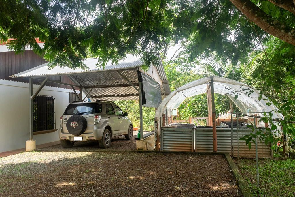 Silver SUV parked under a metal carport beside a curved, plastic-covered greenhouse in a shaded, leafy yard.