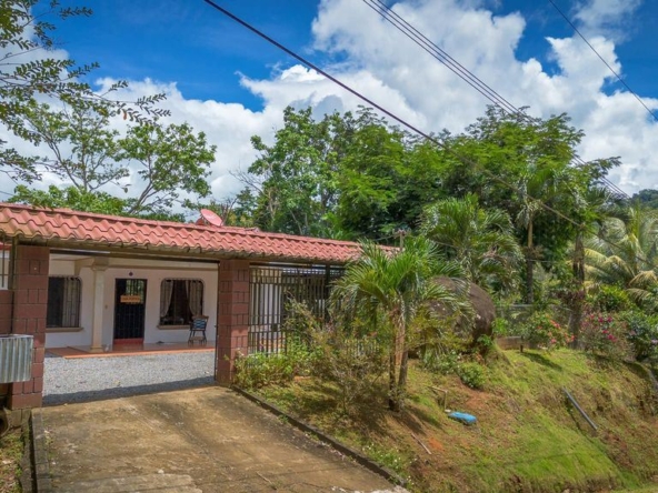 Single-story house with a red-tiled roof and white walls, front porch under a covered veranda, on a sloped gravel driveway surrounded by tropical trees.