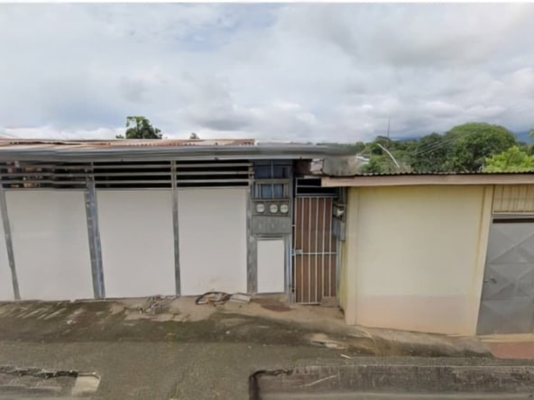 A row of small, grey metal-fronted buildings with gates on a street, weathered walls and a concrete sidewalk under a cloudy sky.