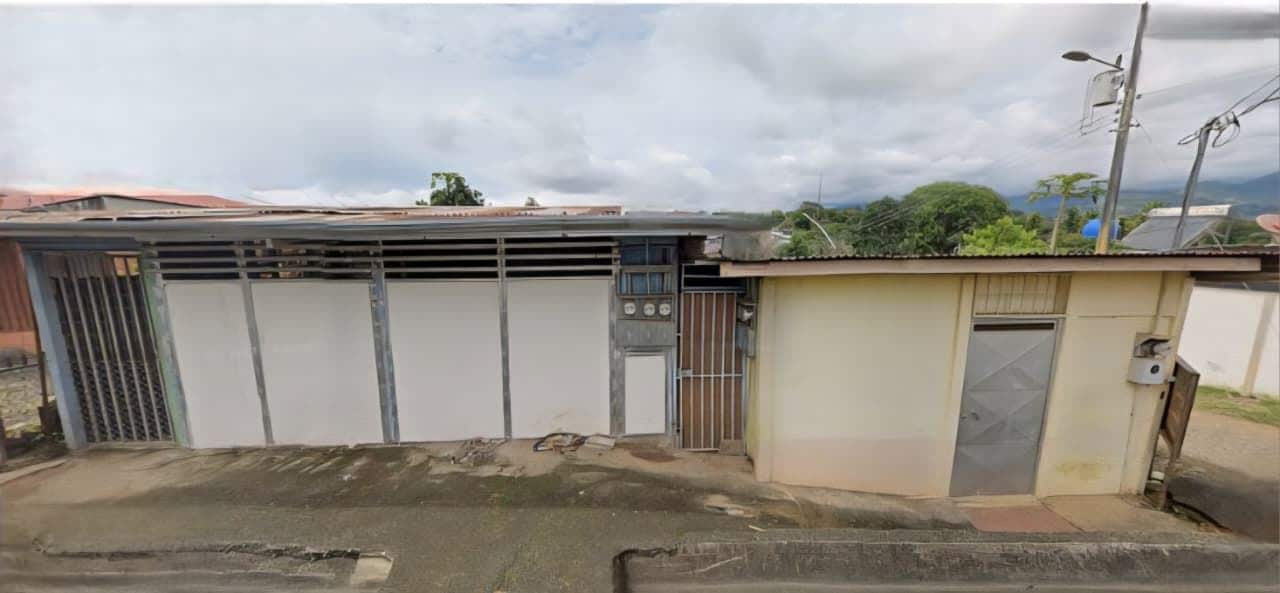 A row of small, grey metal-fronted buildings with gates on a street, weathered walls and a concrete sidewalk under a cloudy sky.