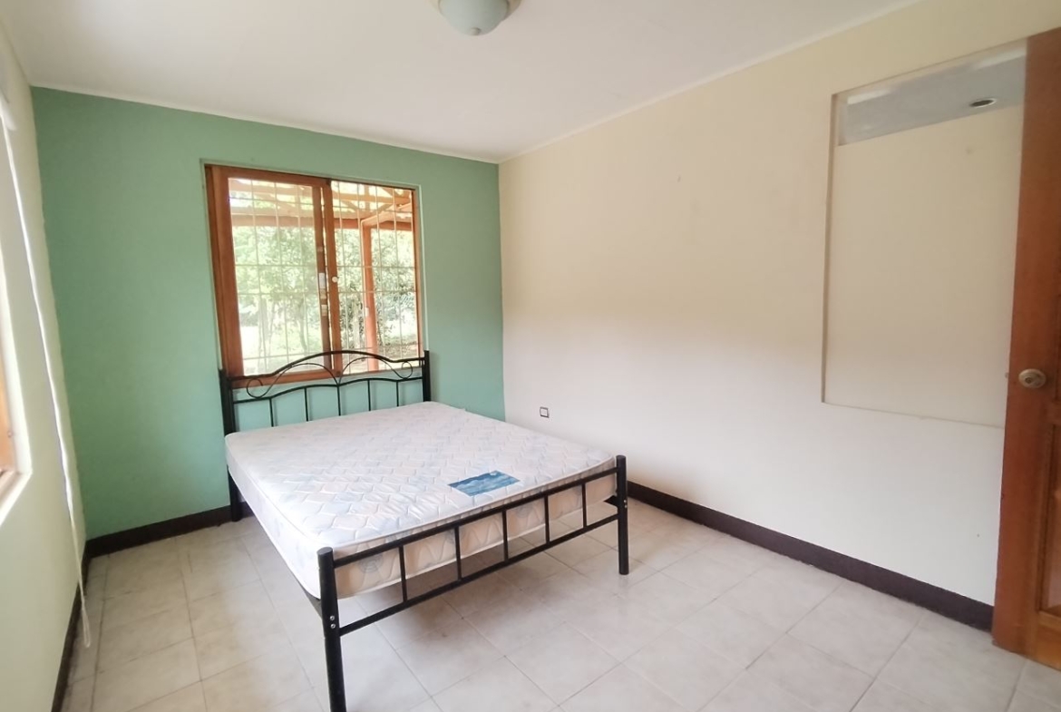 Bedroom with a metal-framed bed and white mattress against a green accent wall by a window with bars on a tiled floor, minimal furniture present.