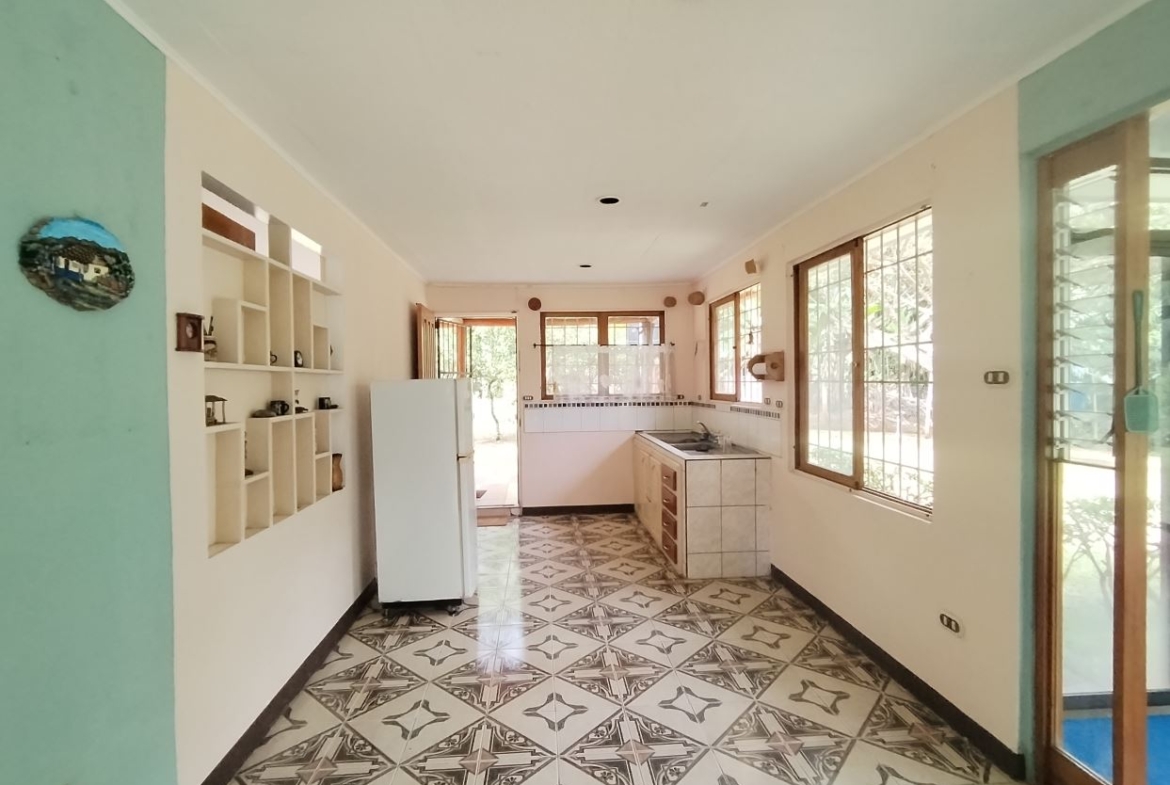 A bright galley-style kitchen with white shelves, a white fridge, tiled counters, and large windows overlooking a garden at the far end.