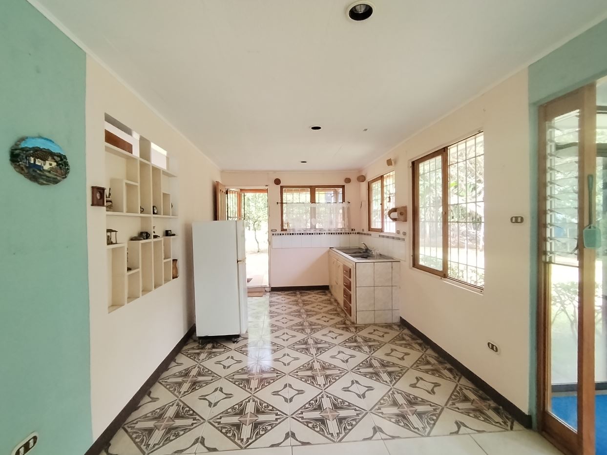 A bright galley-style kitchen with white shelves, a white fridge, tiled counters, and large windows overlooking a garden at the far end.