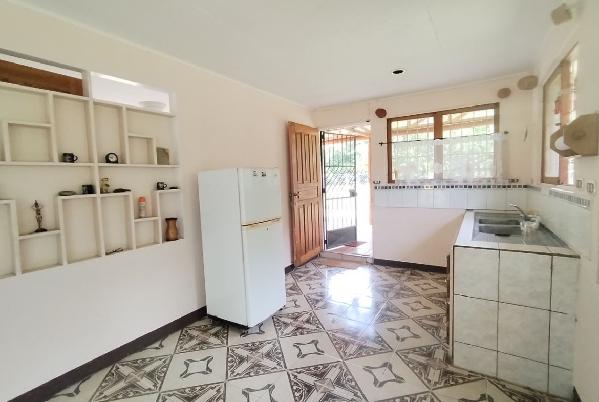 Bright kitchen with a white fridge, tiled counter, sink, and decorative open shelves; door to outside visible.