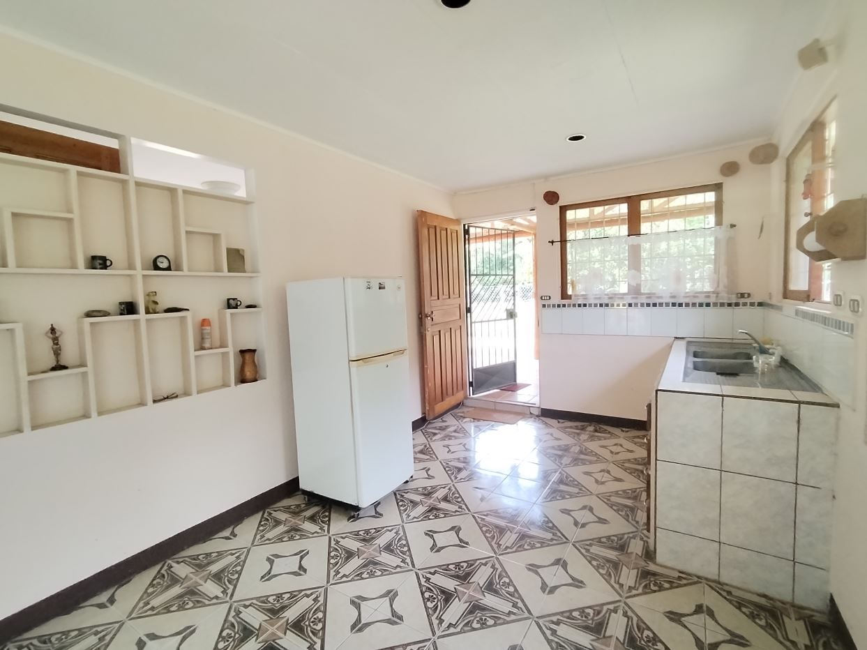 Bright kitchen with a white fridge, tiled counter, sink, and decorative open shelves; door to outside visible.
