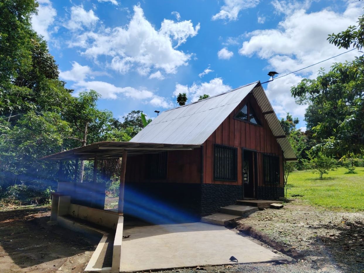 Rustic wooden cabin with a silver metal gable roof and a triangular attic window, set in a green yard under a bright blue sky.