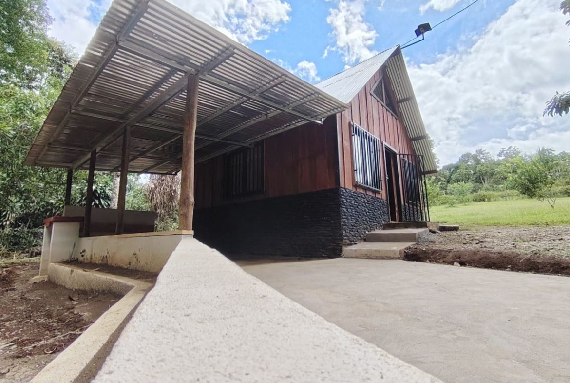 Rural wooden house with a slanted metal roof, stone foundation, and a covered porch on the left among green trees.