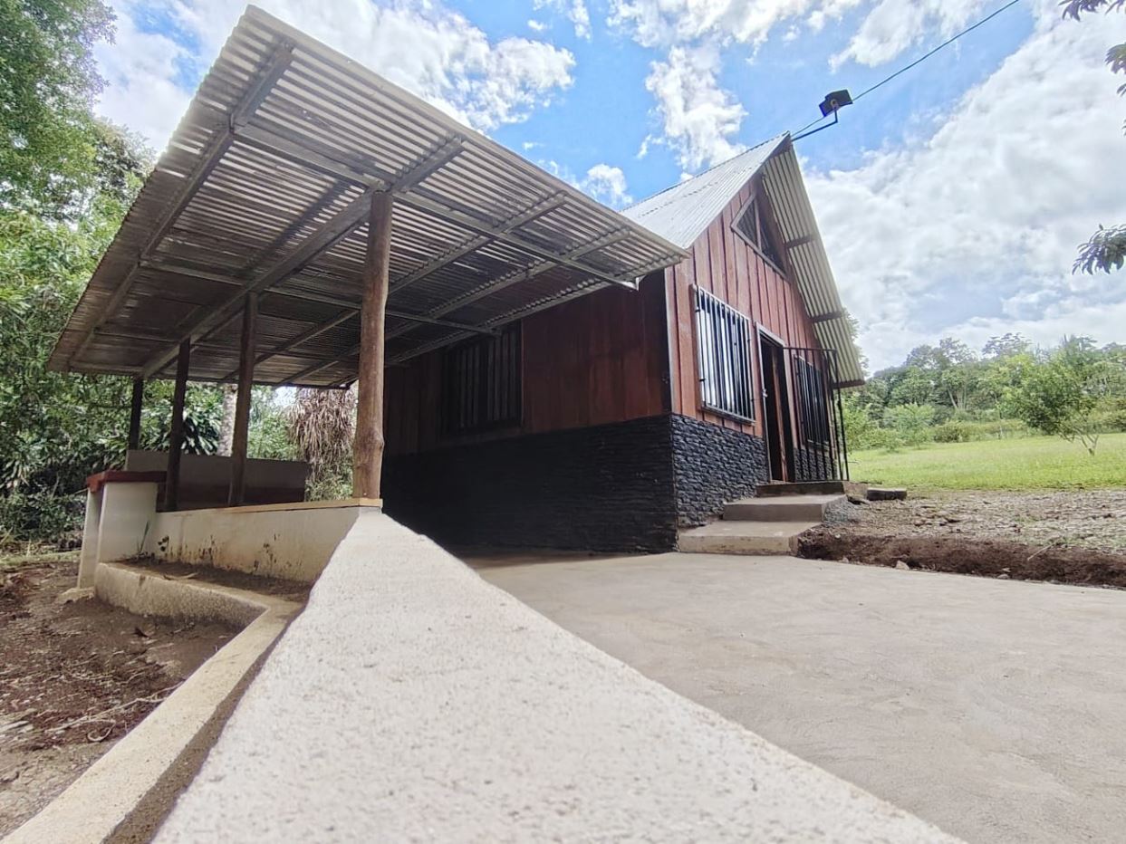 Rural wooden house with a slanted metal roof, stone foundation, and a covered porch on the left among green trees.
