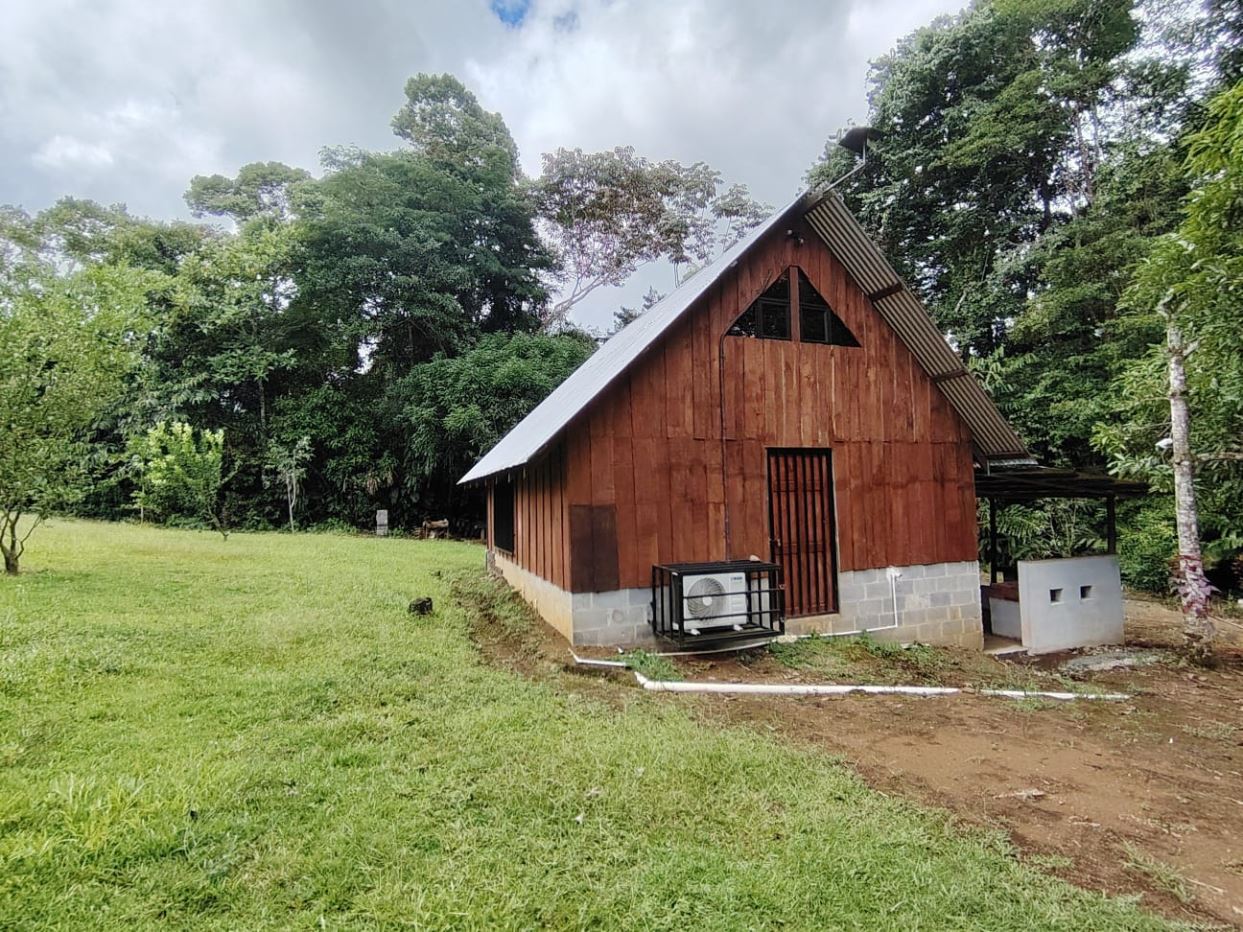 Small wooden cabin with a steep metal roof in a grassy clearing, surrounded by trees.