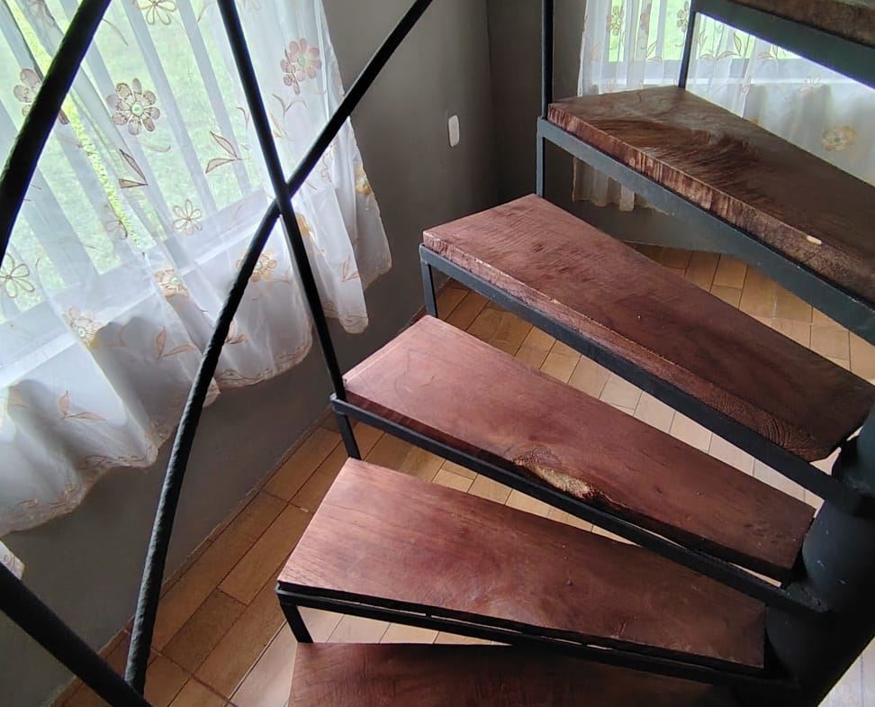Spiral staircase with dark metal railing and wooden steps beside a window with sheer floral curtains.