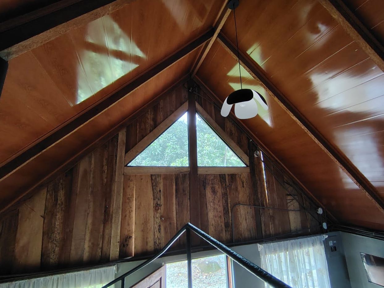 A rustic attic interior with dark wood walls, exposed beams, and a triangular window at the peak letting in greenery. A modern white-and-black pendant light hangs from the sloped ceiling above a railing.