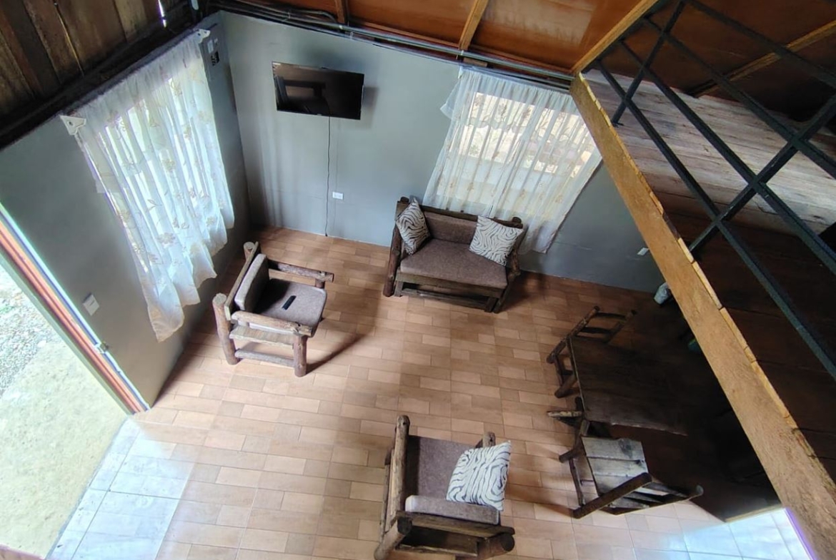 Bird's-eye view of a rustic living room with wooden armchairs, a two-seat sofa, wall-mounted TV, and a tiled floor.