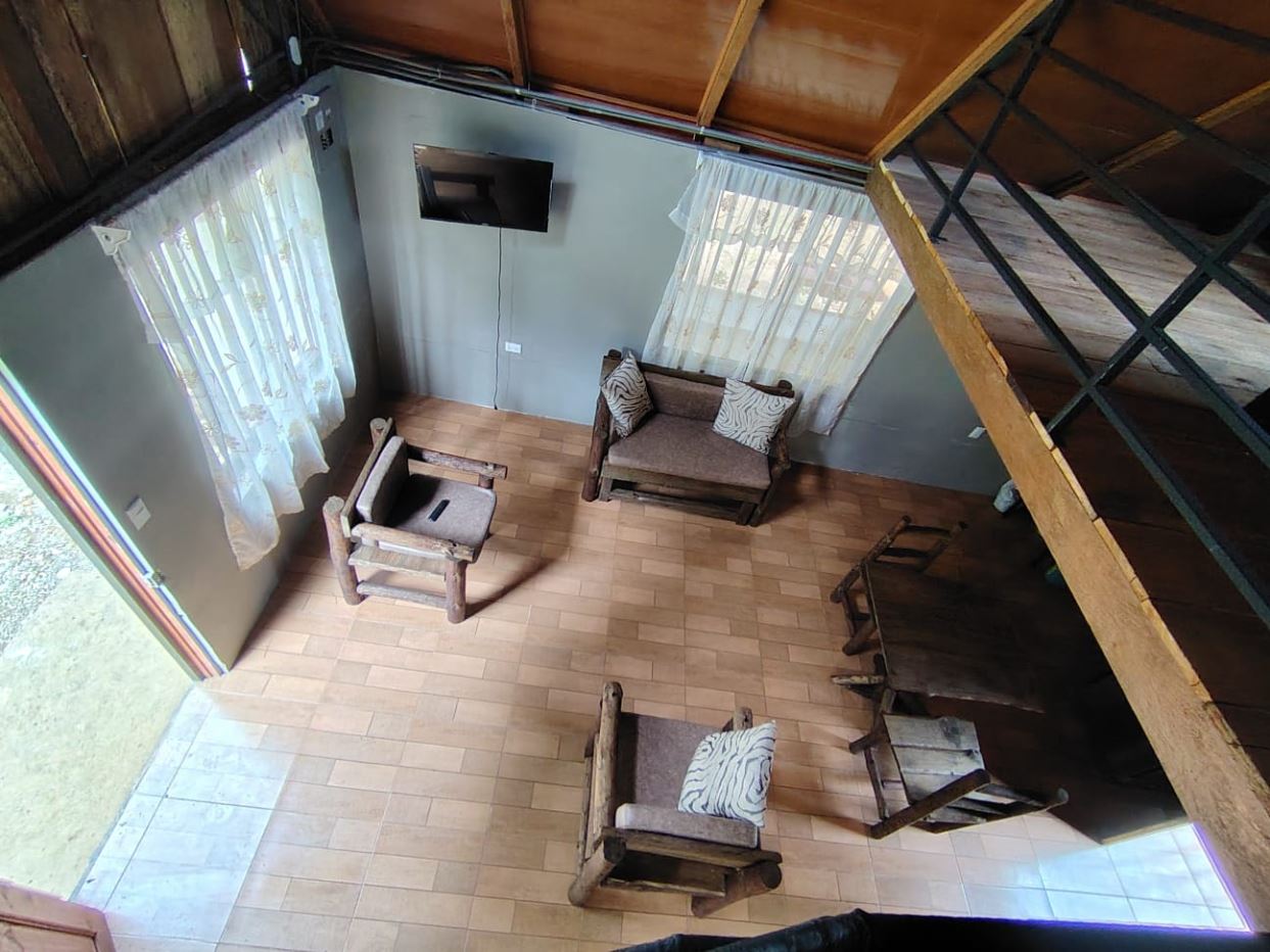 Bird's-eye view of a rustic living room with wooden armchairs, a two-seat sofa, wall-mounted TV, and a tiled floor.
