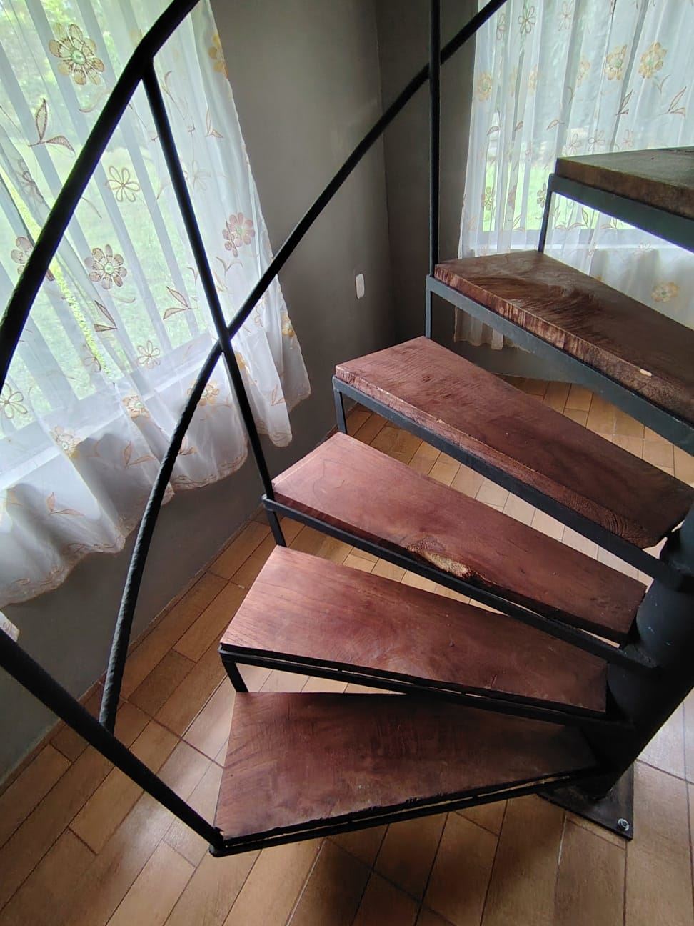 Spiral staircase with dark metal railing and wooden steps beside a window with sheer floral curtains.