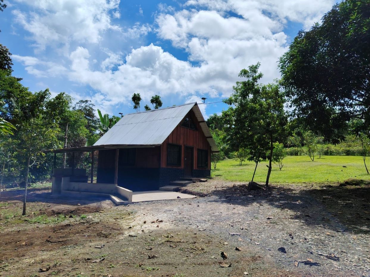 Small rustic cabin with a metal gabled roof, wooden siding, and a covered porch set in a grassy, tree-filled yard under a blue sky with clouds.