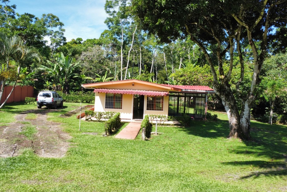 Small beige cottage with a red awning in a grassy yard, a white SUV parked to the left and tall tropical trees surrounding the scene.