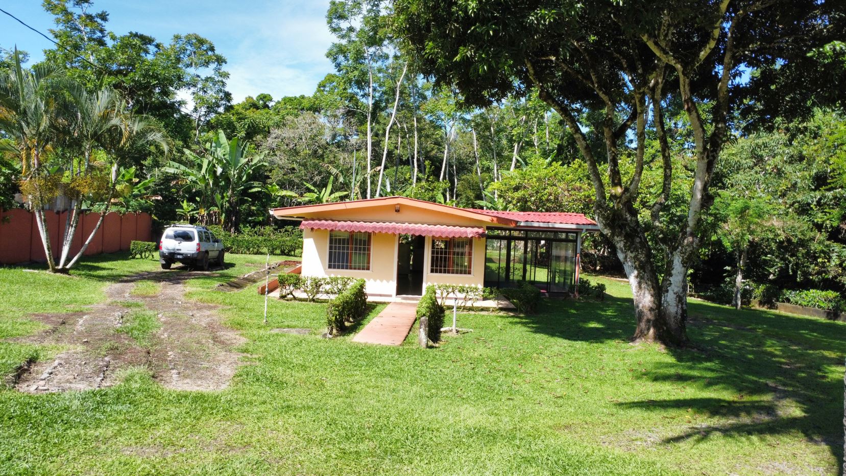 Small beige cottage with a red awning in a grassy yard, a white SUV parked to the left and tall tropical trees surrounding the scene.