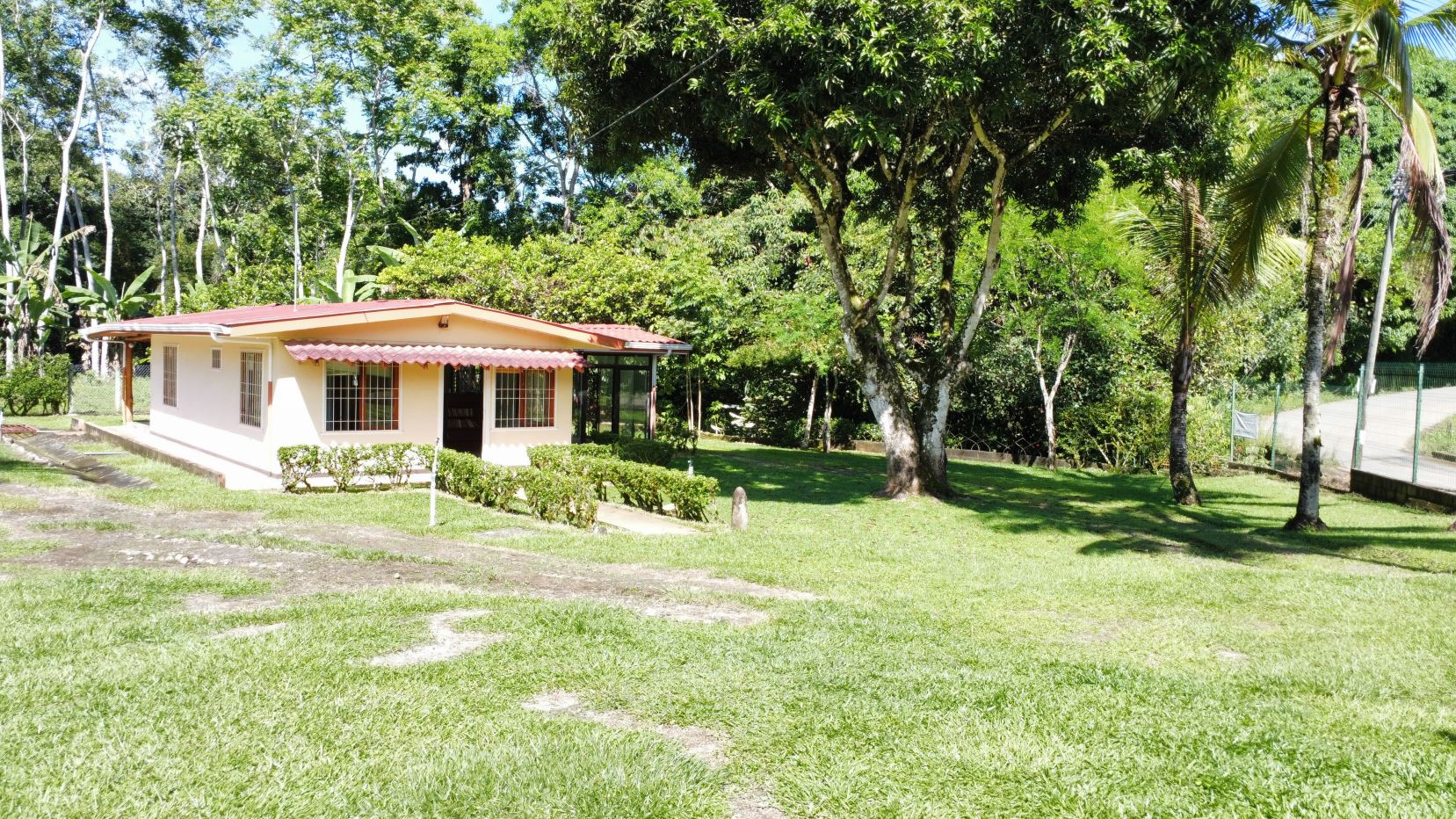 Single-story cream house with a red awning, black-framed windows, and a small hedge, set in a sunny green yard with trees nearby