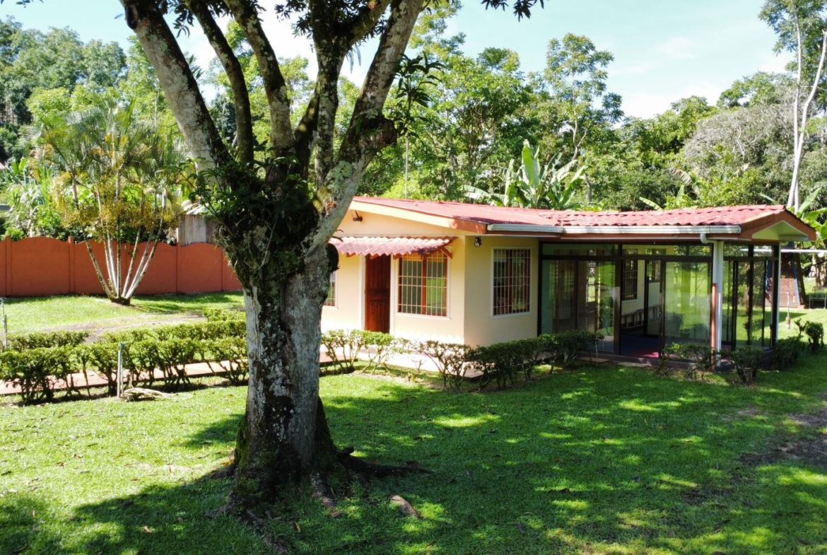 Single-story beige house with a red tiled roof and glass-walled sunroom, set in a lush tropical yard with a large tree in the foreground.