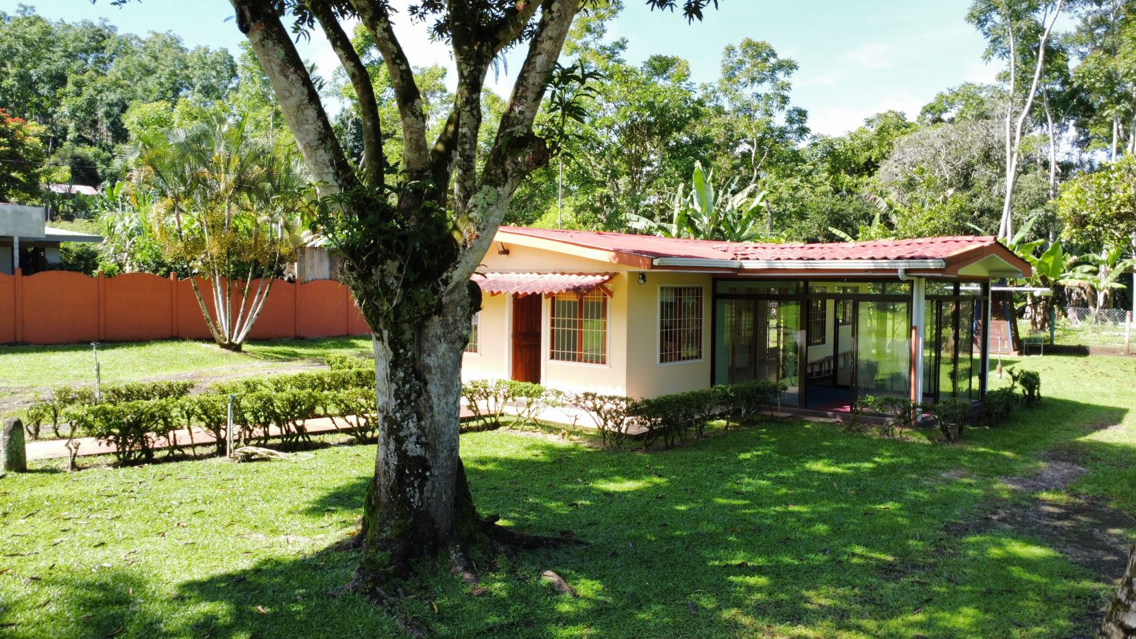 Single-story beige house with a red tiled roof and glass-walled sunroom, set in a lush tropical yard with a large tree in the foreground.