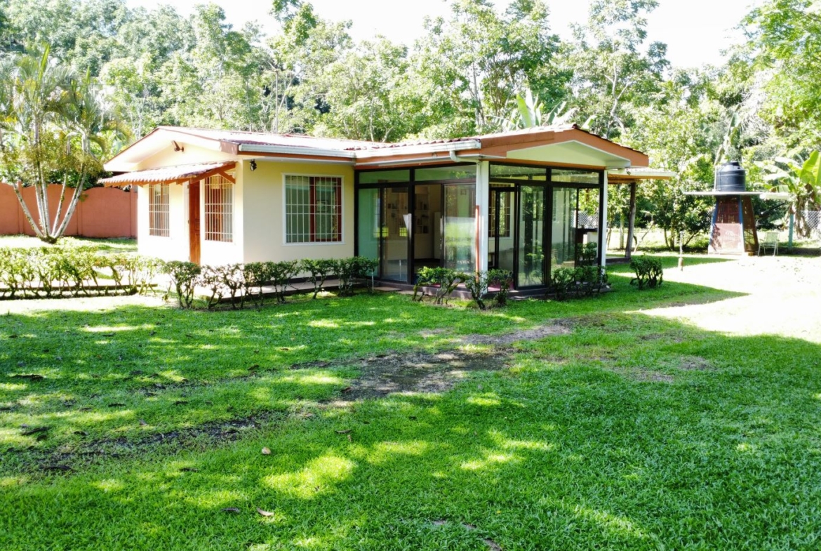 Single-story house with a glass-walled sunroom, beige exterior, and a green lawn surrounded by trees and hedges.