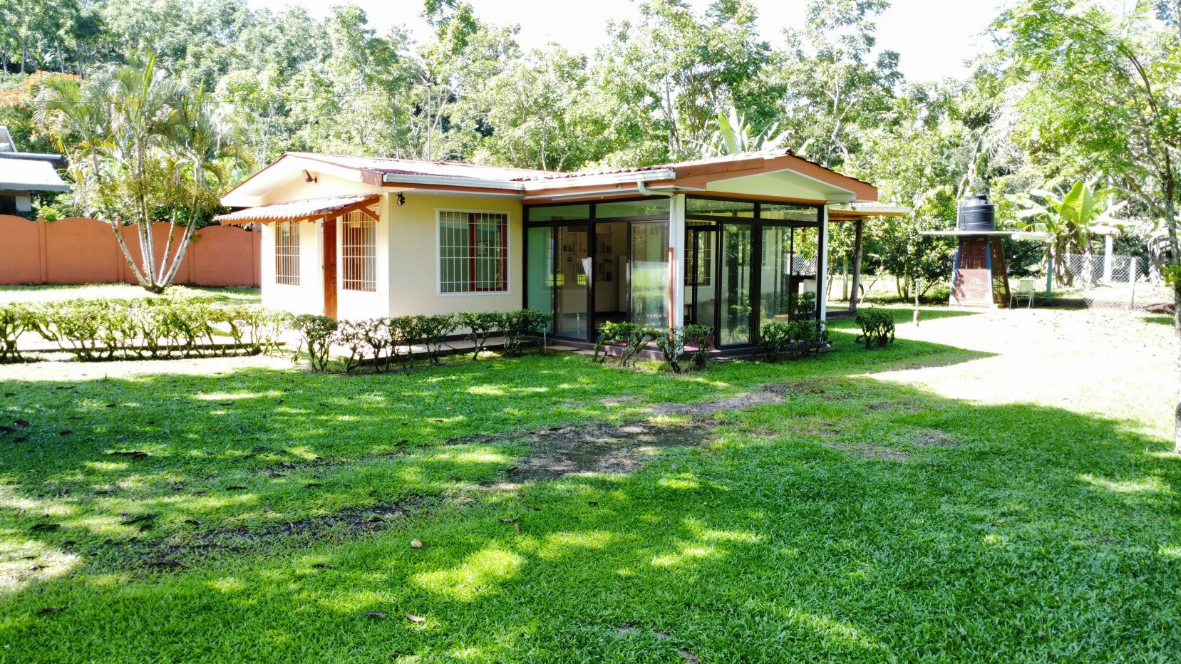 Single-story house with a glass-walled sunroom, beige exterior, and a green lawn surrounded by trees and hedges.