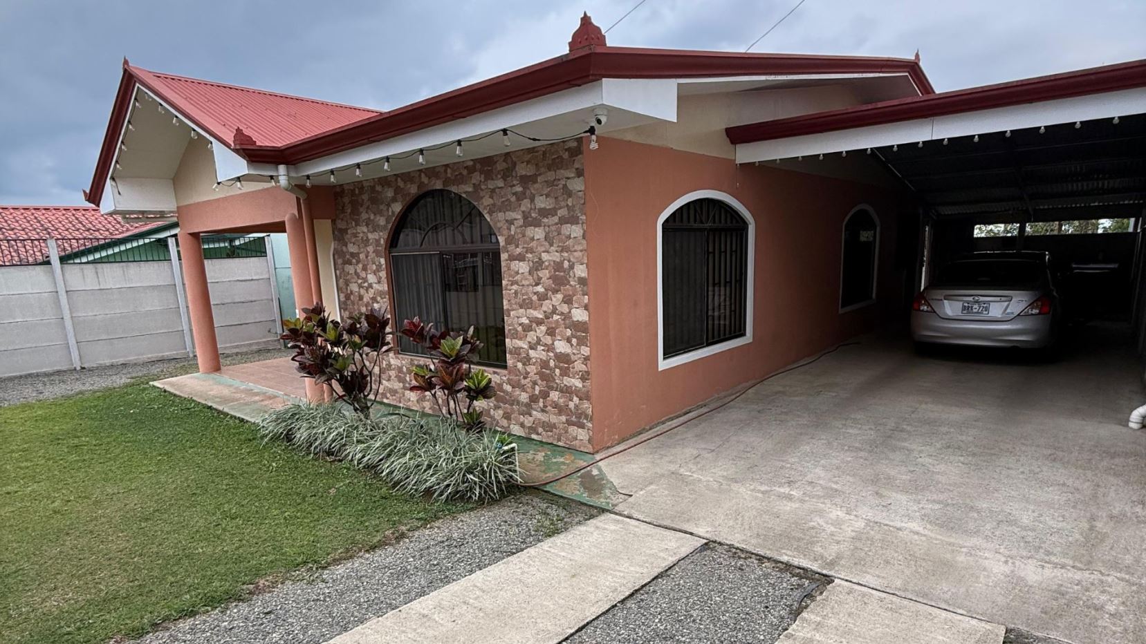 Single-story house with a covered carport on the right, a parked silver car, and a stone-accent wall by two arched windows; a small garden bed with tropical plants in front.