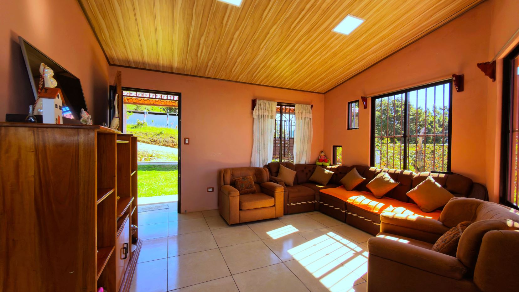 Sunlit living room with warm orange walls, a wooden ceiling, and a large L-shaped brown sofa with many cushions facing windows and a garden outside.