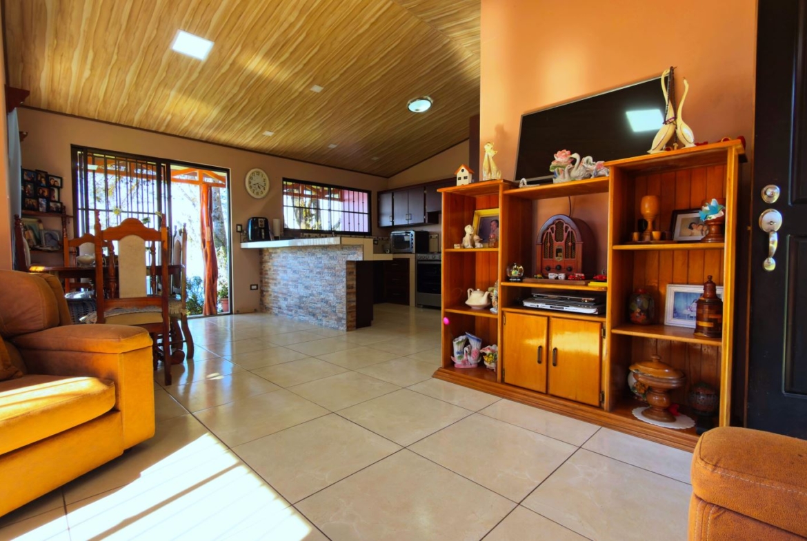 Sunny living room with orange walls, wooden ceiling, and tiled floor; open kitchen with stone island in the background and a dining area to the left.