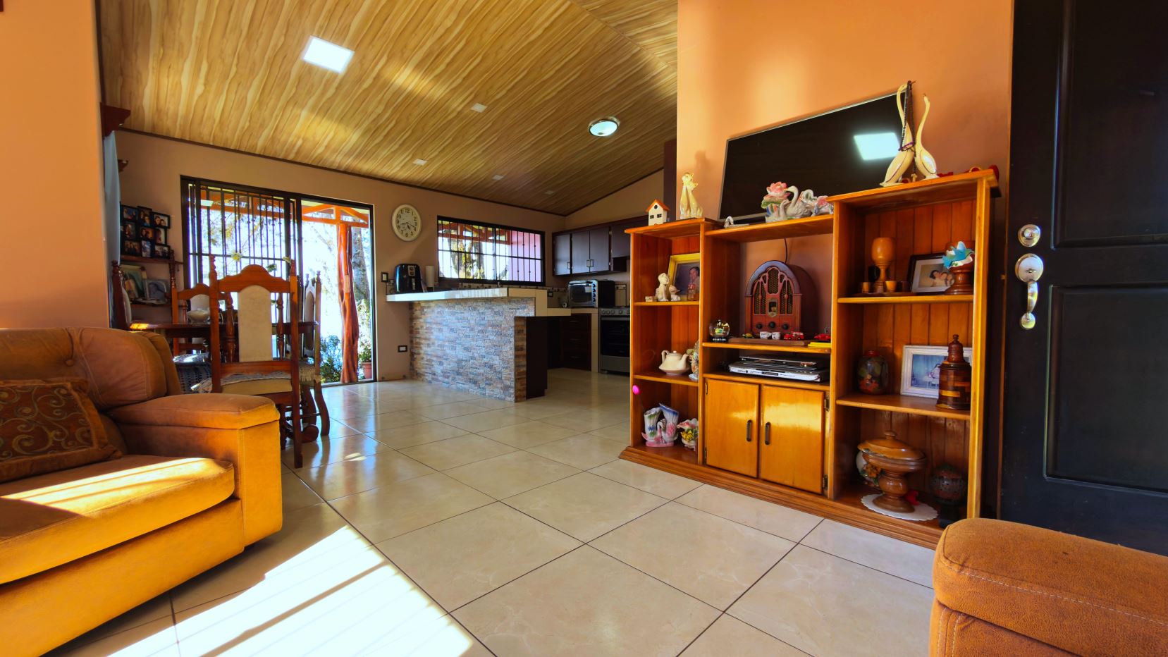 Sunny living room with orange walls, wooden ceiling, and tiled floor; open kitchen with stone island in the background and a dining area to the left.