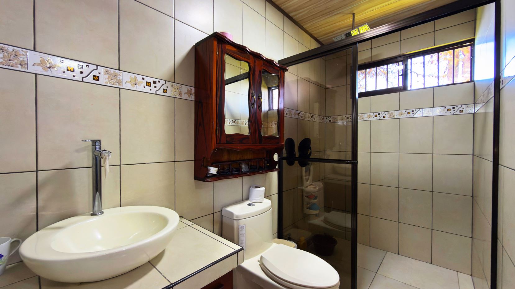 Beige-tiled bathroom with a white vessel sink, chrome faucet, wooden mirrored cabinet, toilet, and glass shower enclosure.