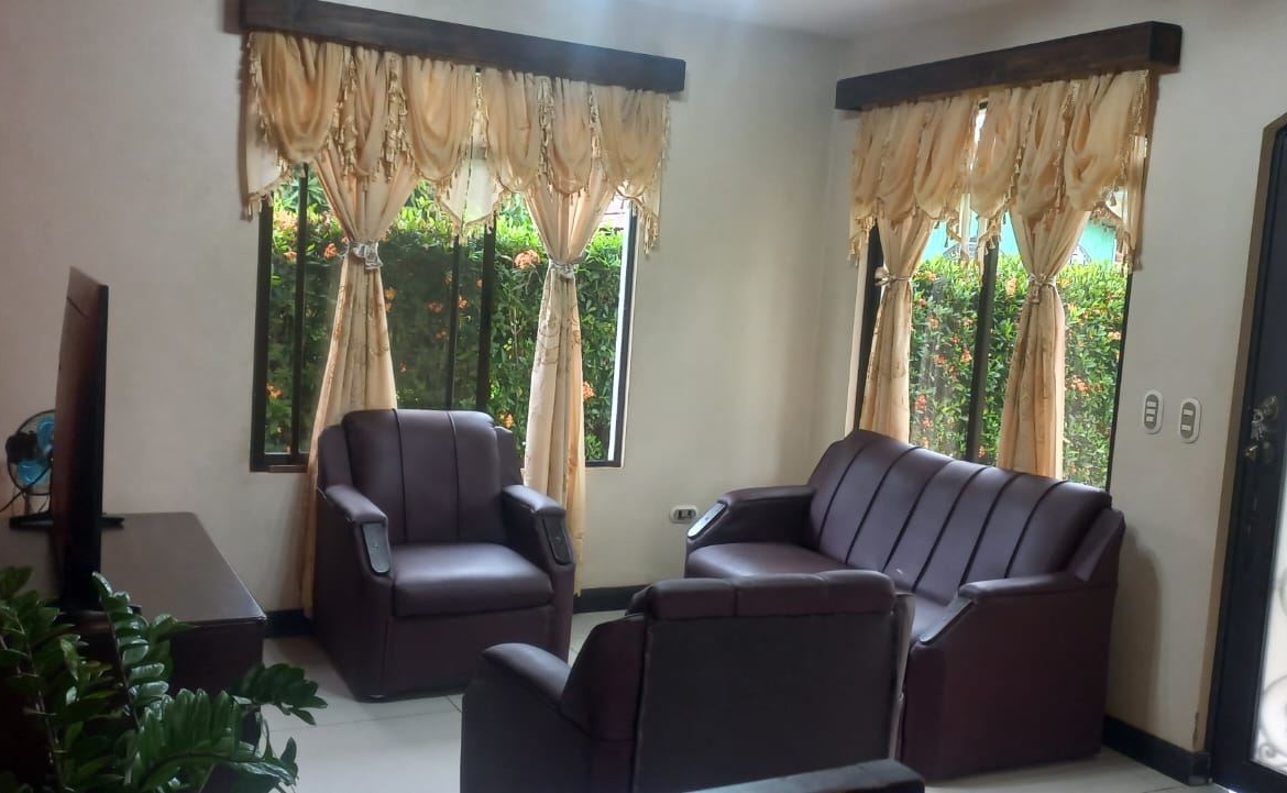 Cozy living room with brown leather sofa set around a TV, beige draped windows and a view of greenery outside the windows.