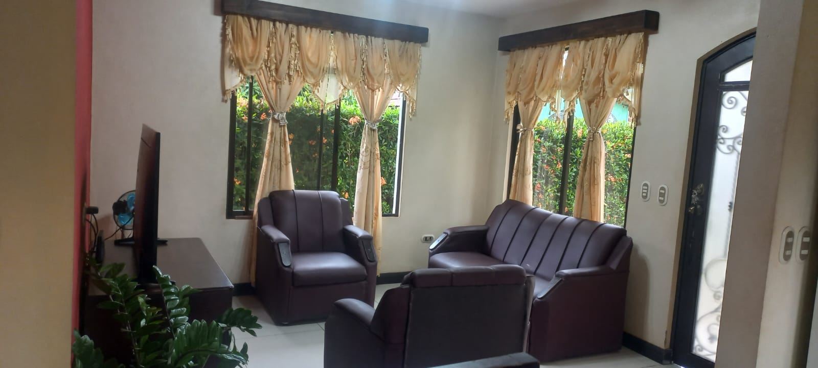Cozy living room with brown leather sofa set around a TV, beige draped windows and a view of greenery outside the windows.