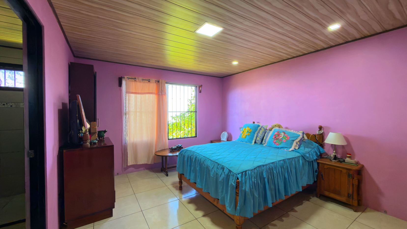 Bedroom with pink walls and blue bedspread; window with sheer curtains beside a wooden bed.