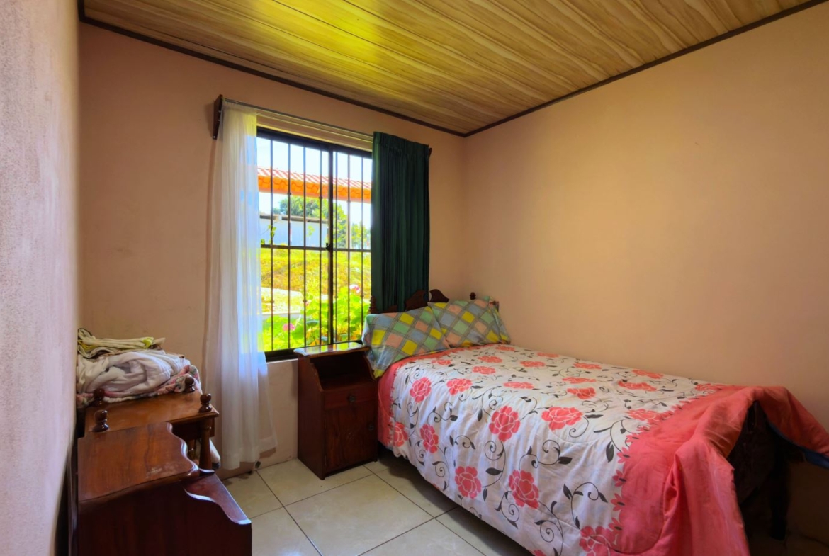 A small bedroom with a single bed covered in a pink floral duvet, wooden nightstand, and a window with bars and sheer white curtains, overlooking a sunny green yard.