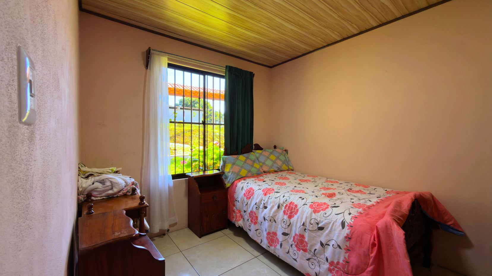 A small bedroom with a single bed covered in a pink floral duvet, wooden nightstand, and a window with bars and sheer white curtains, overlooking a sunny green yard.
