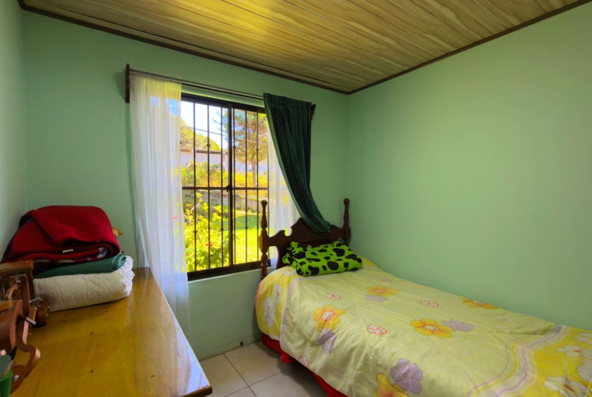 Cozy bedroom with pale green walls, a single bed with yellow floral sheets, and a window with white sheer and dark green curtains.