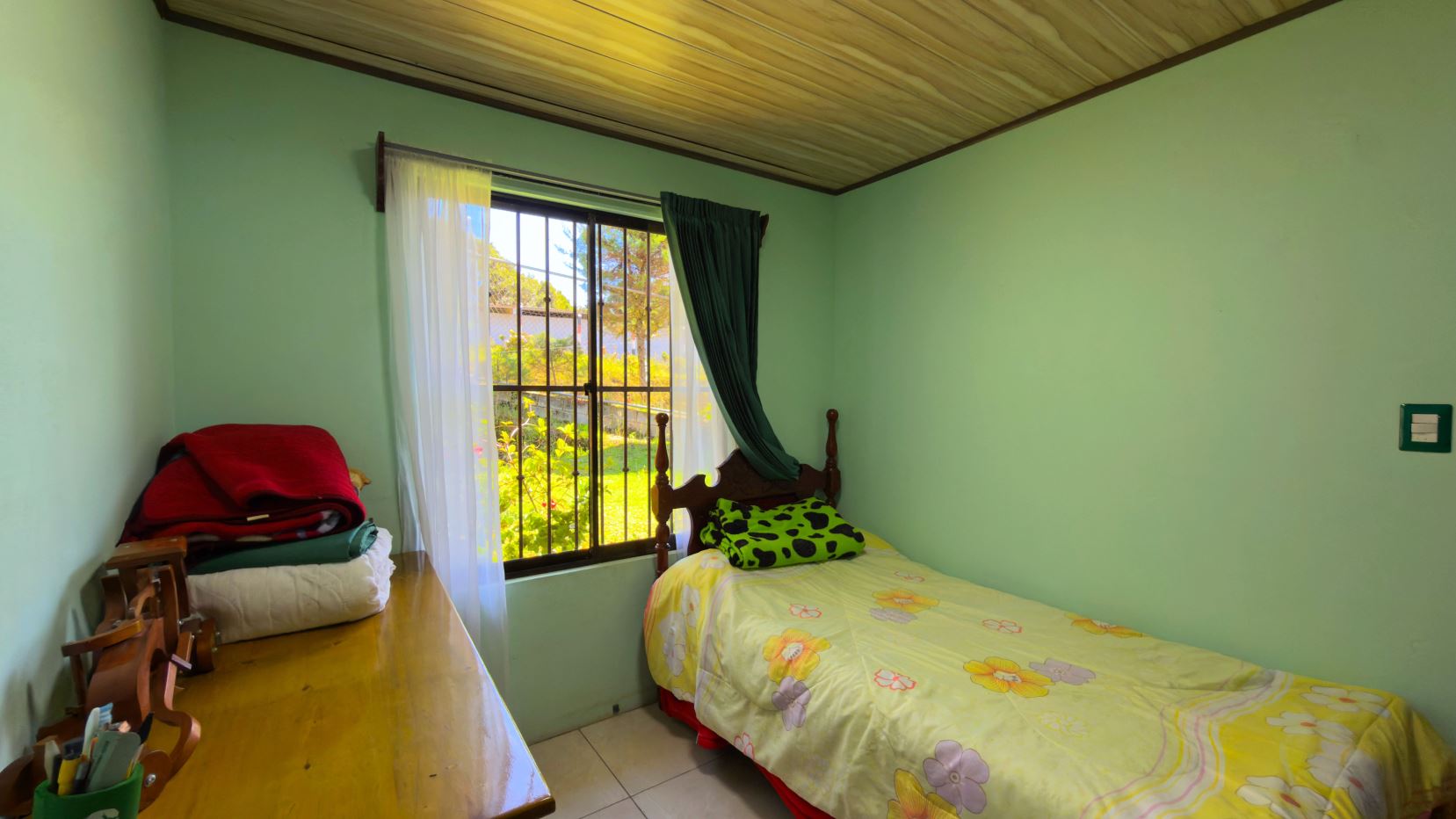 Cozy bedroom with pale green walls, a single bed with yellow floral sheets, and a window with white sheer and dark green curtains.