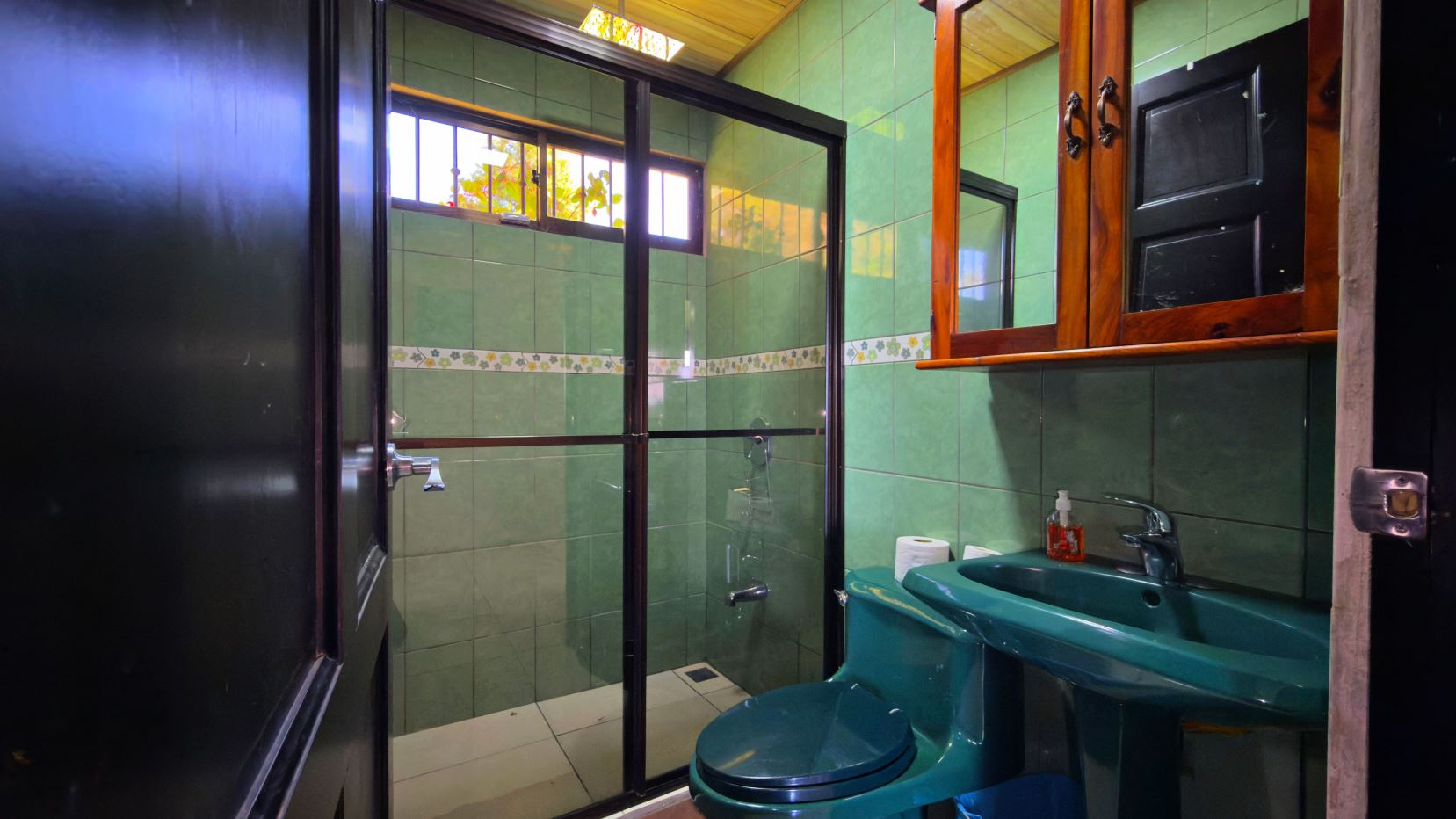 Green tiled bathroom with a glass-walled shower, and a matching green toilet and sink; wooden cabinet above the sink.