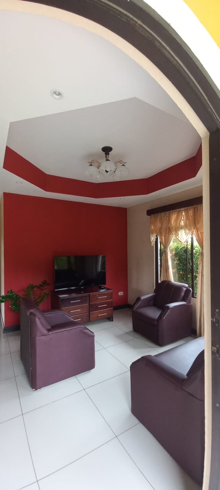 Living room with red accent wall, white tile floor, and four purple armchairs around a wooden TV console with a flat-screen TV.