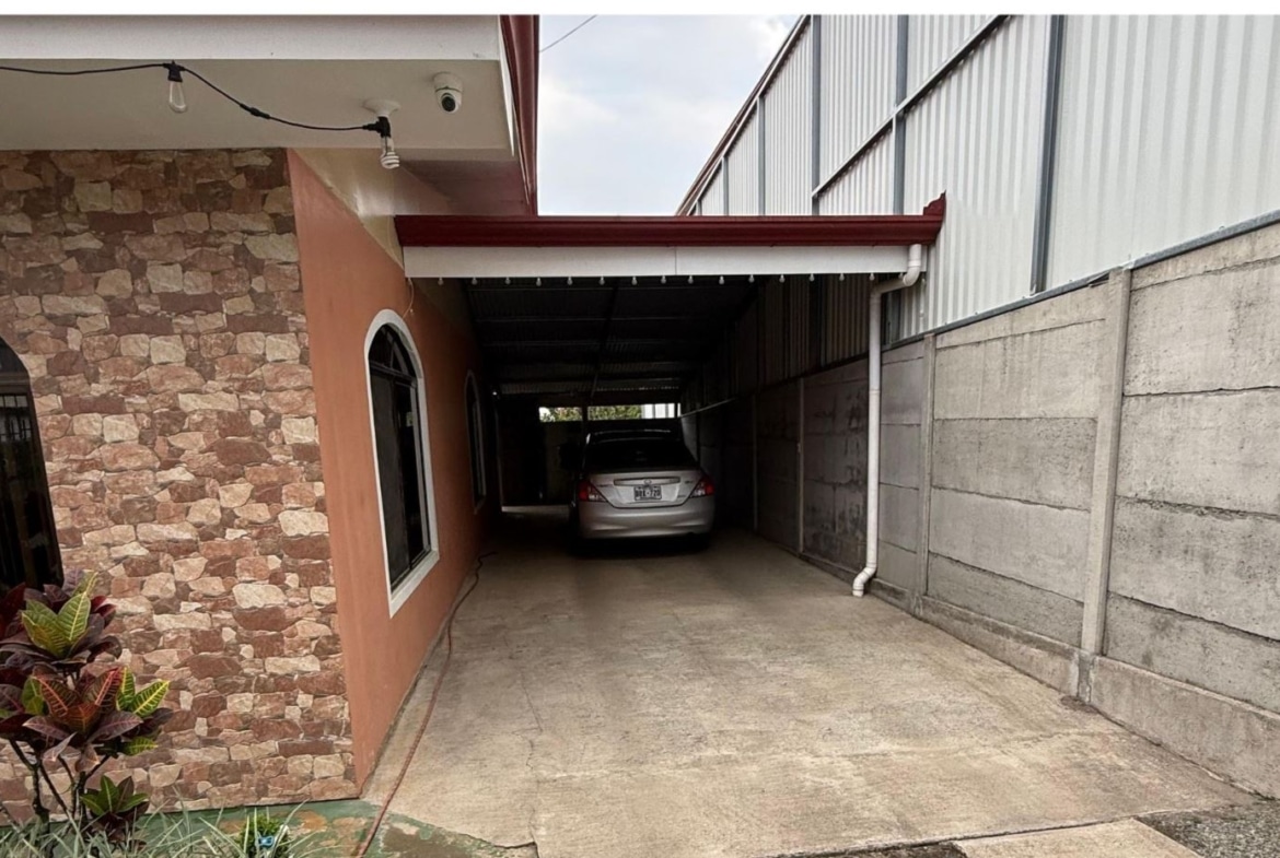 Carport with a silver sedan parked at the end, between a stone-patterned wall on the left and a concrete wall on the right, under string lights.