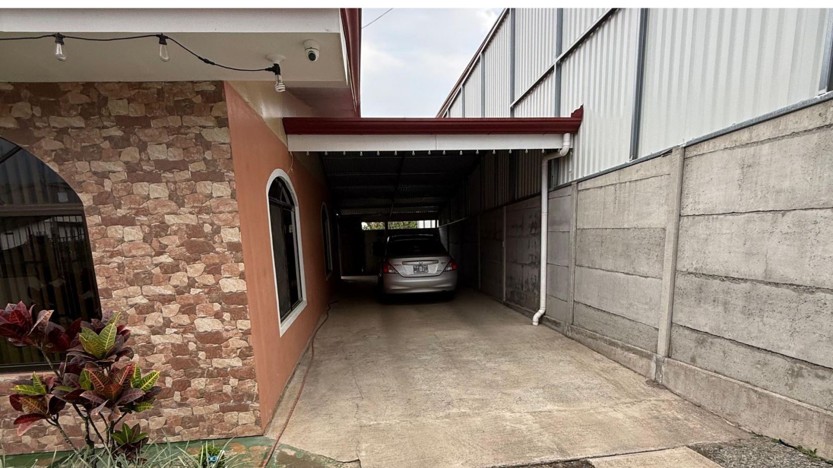 Carport with a silver sedan parked at the end, between a stone-patterned wall on the left and a concrete wall on the right, under string lights.
