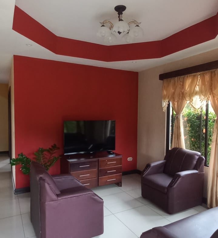Living room with a red accent wall, a flat-screen TV on a wooden console, and burgundy leather chairs by a large window with beige curtains.